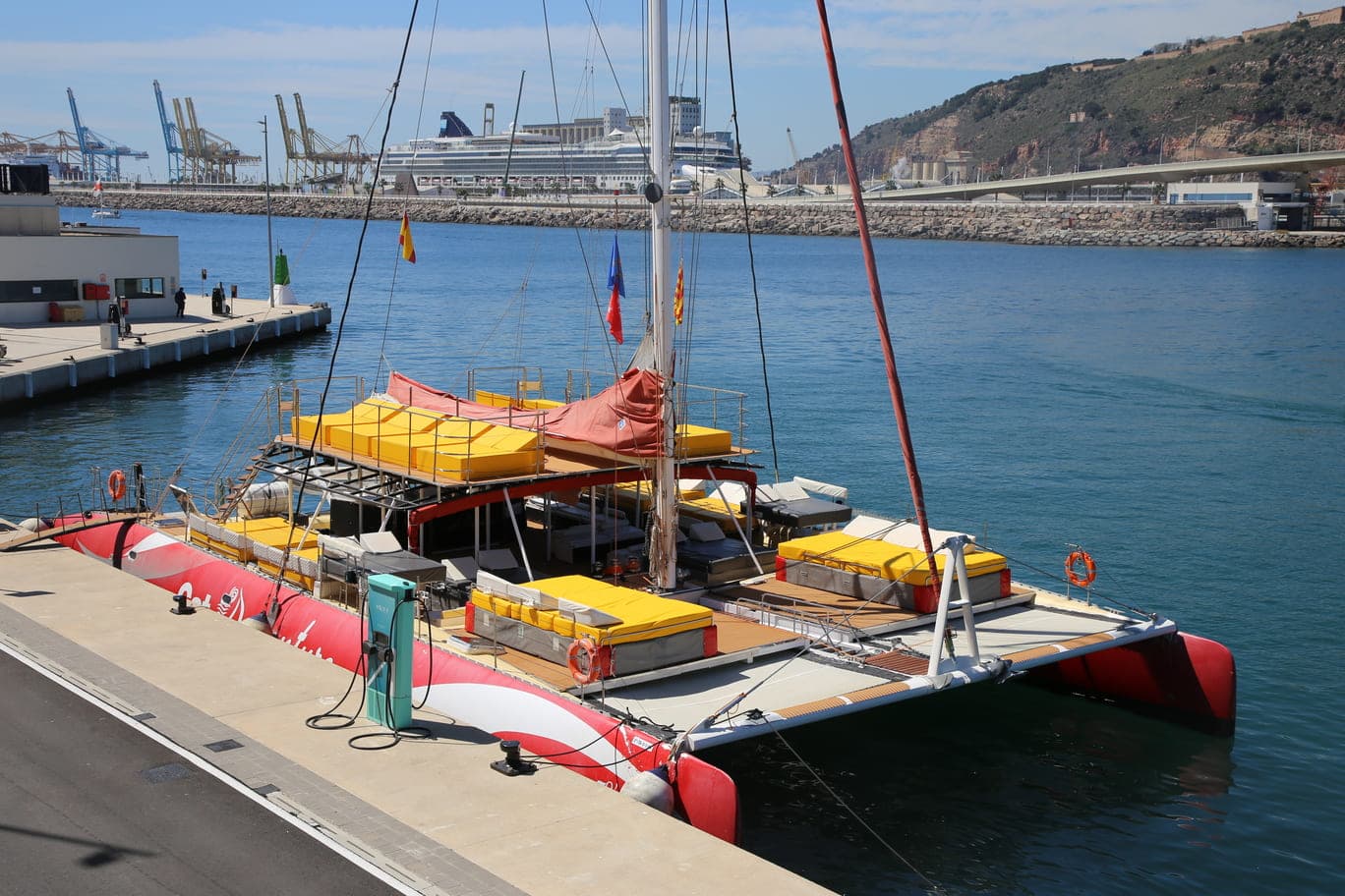 Vista elevada del catamarán rojo amarrado en el puerto con grúas y crucero al fondo