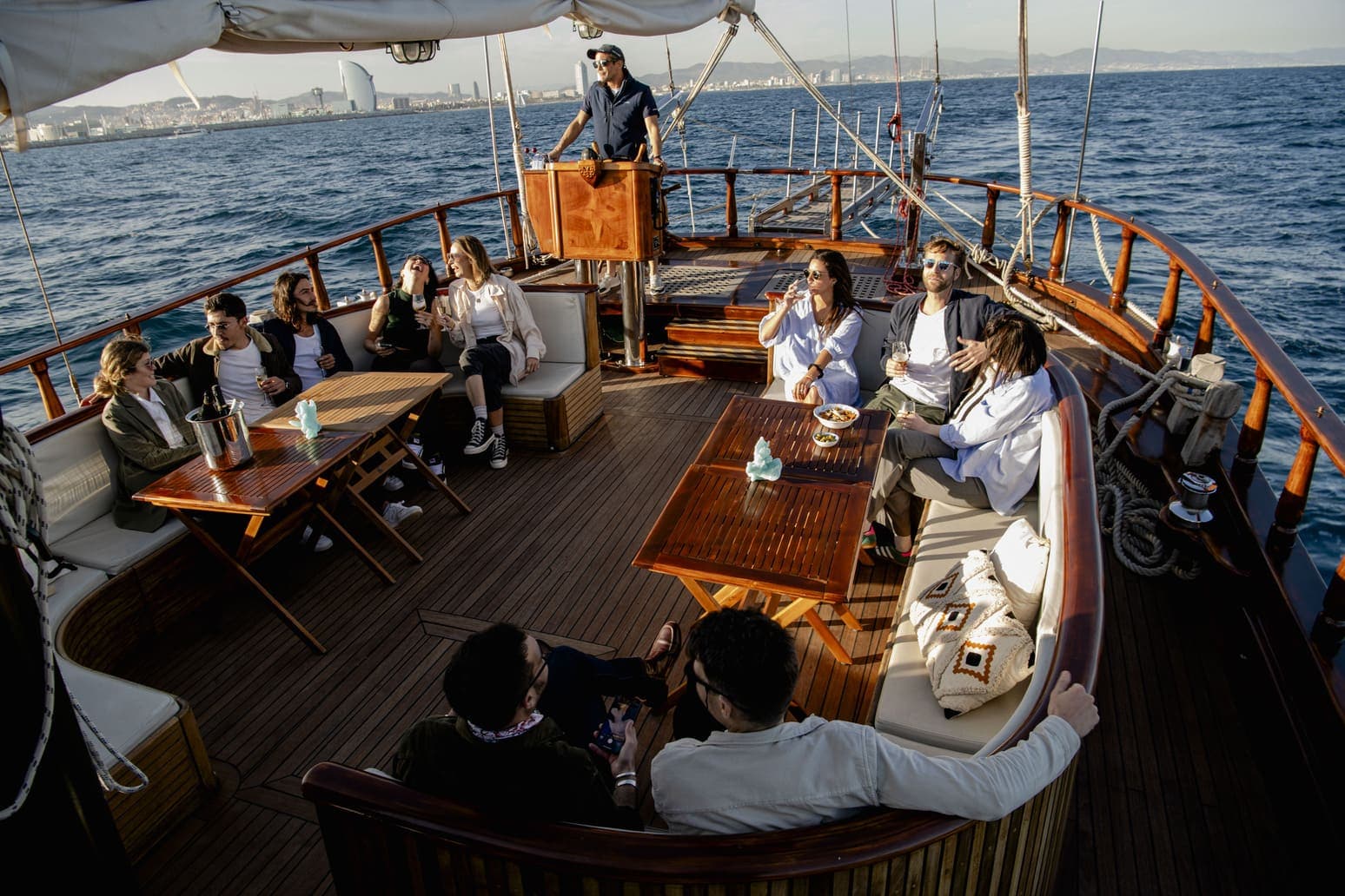 Grupo de personas en la cubierta de un velero de madera con el capitán al timón y el hotel W al fondo