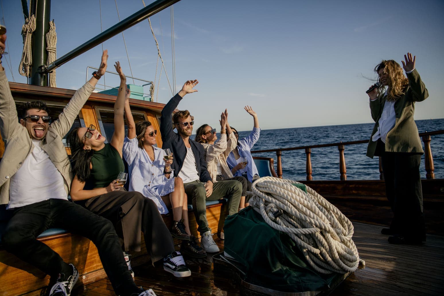 Grupo de amigos brindando y bailando en la cubierta de un velero mientras una mujer canta con micrófono