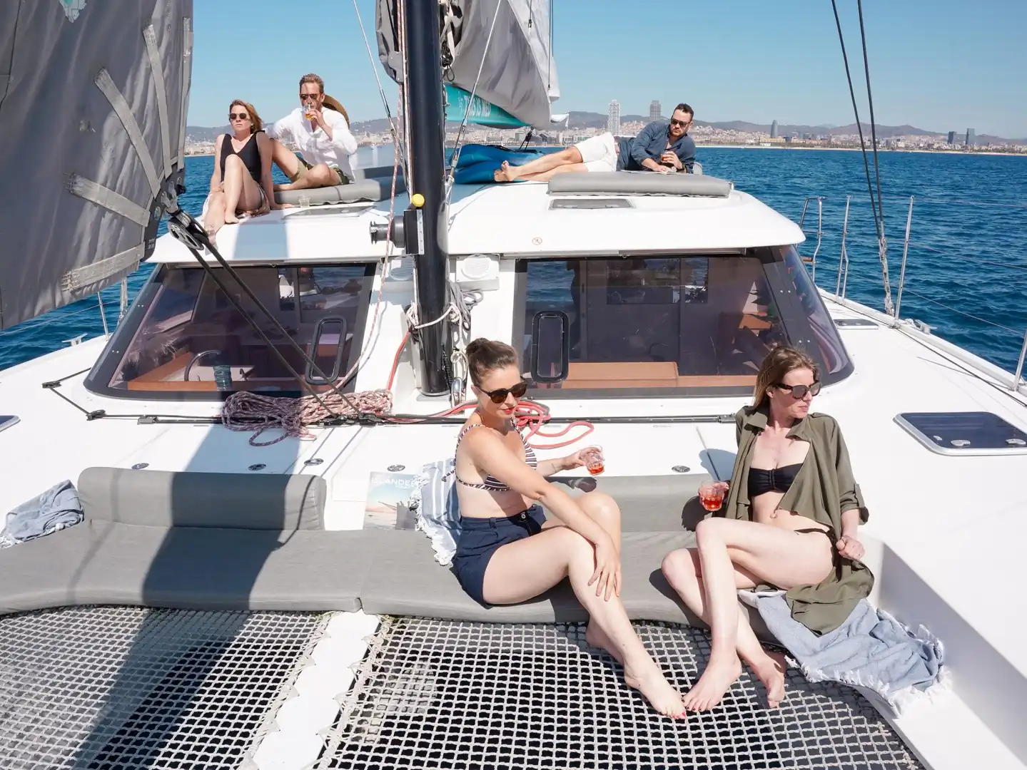 Amigos relajándose con bebidas en la cubierta de un catamarán con el skyline de Barcelona detrás