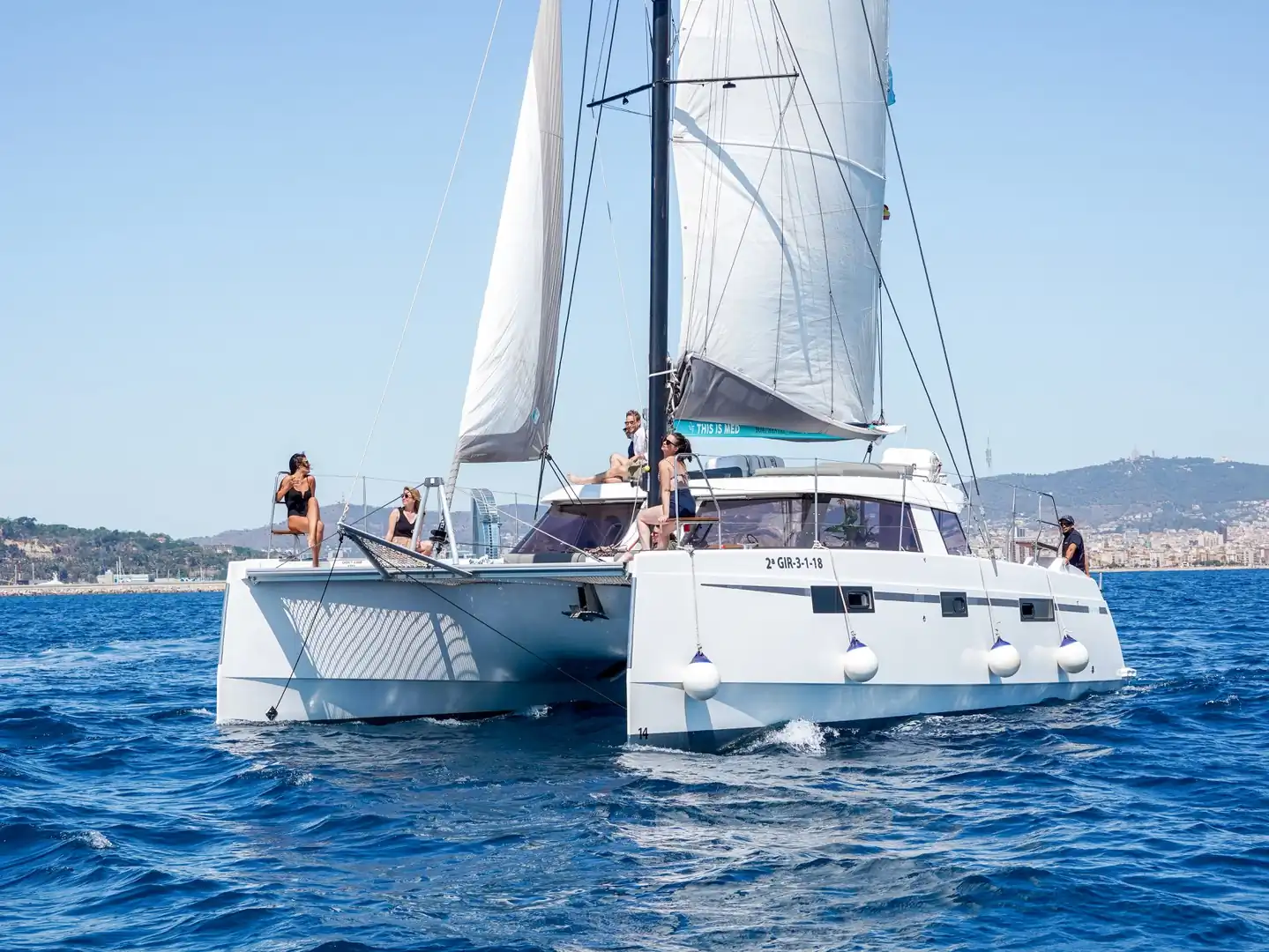Catamarán de vela con pasajeros descansando en la proa y el skyline de Barcelona al fondo
