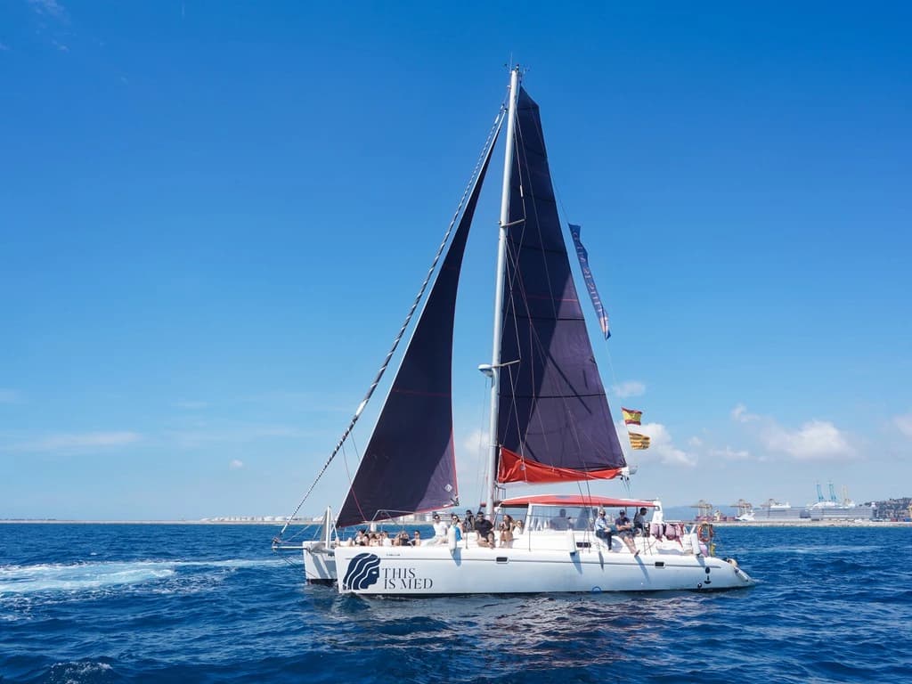 Catamarán navegando con velas oscuras y el logo This is Med en el casco, con grupo a bordo