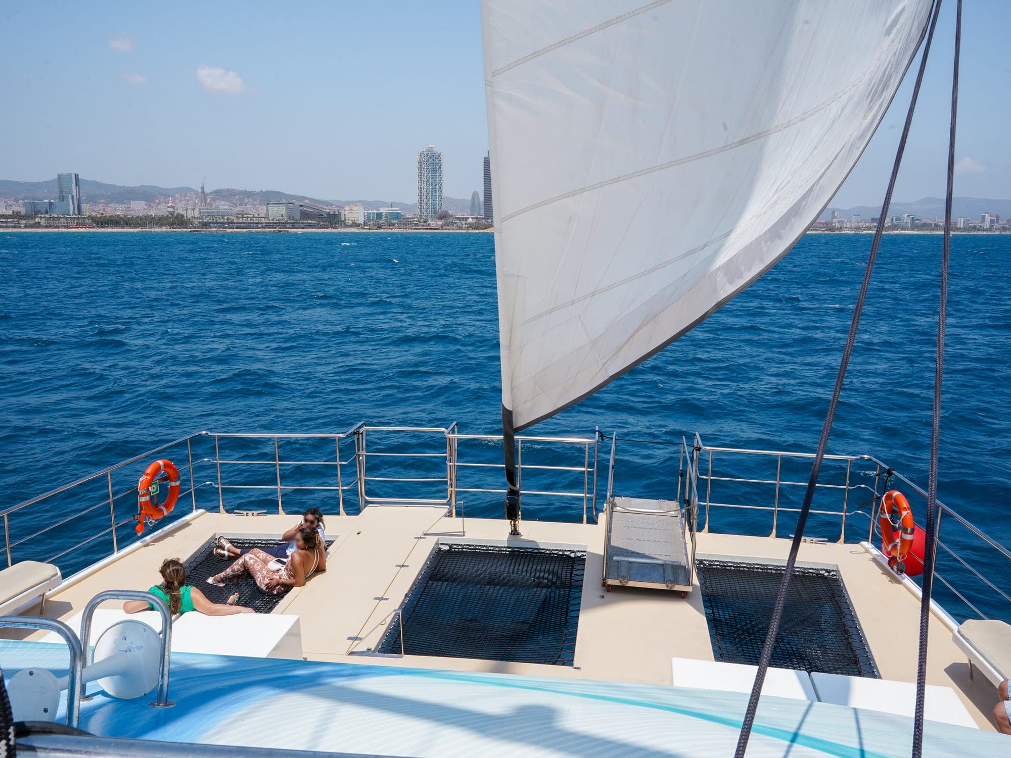 Proa del catamarán con pasajeros relajados navegando frente al skyline de Barcelona