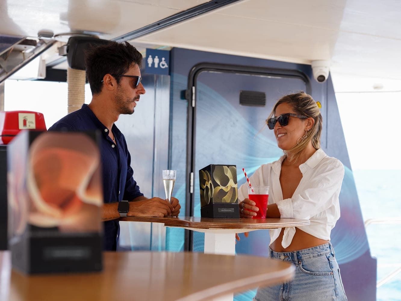 Pareja brindando con cócteles junto a una caja de regalo en la cubierta de un catamarán
